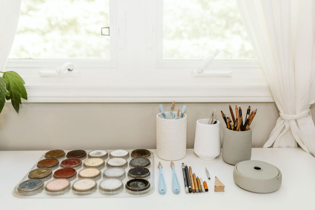 Art supplies including pan pastel palettes, pastel pancils, and containers on a light surface with a window in the background.