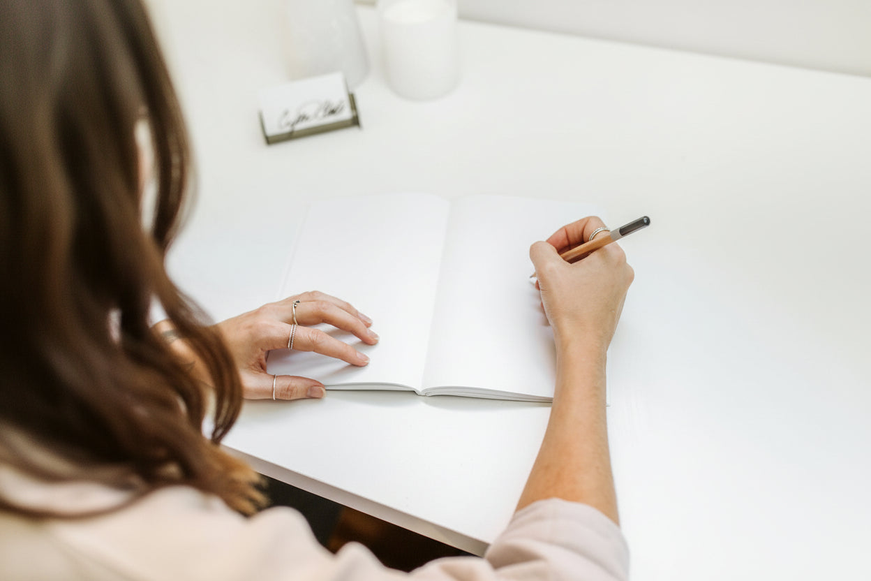 Person writing in a notebook with a pen on a white desk.