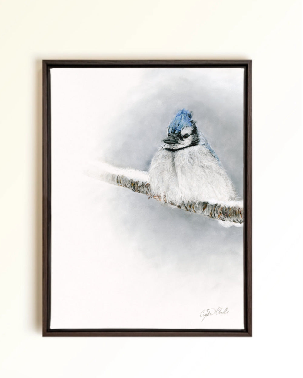 A framed pastel drawing of a blue jay perched on a snowy branch, set against a soft white and blue winter background. The artwork is presented in a dark brown wooden frame and hangs on a light beige wall.