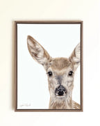 A framed art print featuring a detailed drawing of a young deer fawn with large ears and dark eyes on a white background, enclosed in a brown wooden frame and mounted on a light beige wall.