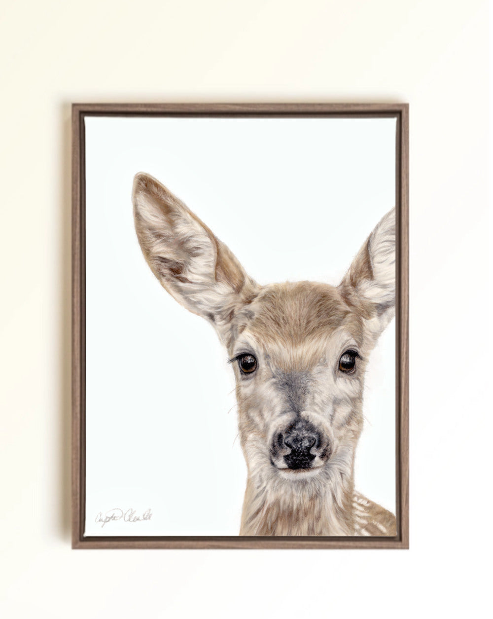 A framed art print featuring a detailed drawing of a young deer fawn with large ears and dark eyes on a white background, enclosed in a brown wooden frame and mounted on a light beige wall.