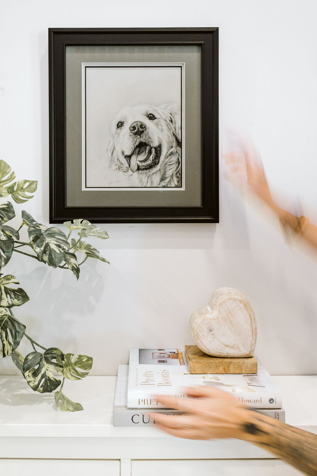 Framed black and white dog portrait on a shelf with books and a plant.