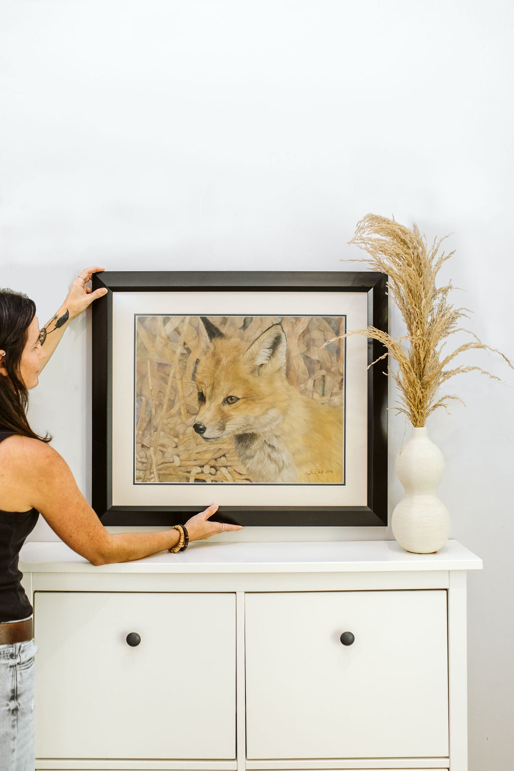 Artist Crystal Clarke adjusting a framed picture of a fox on a white cabinet with decorative elements.