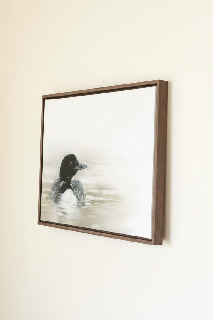 A framed pastel artwork of a loon duck floating on calm water, displayed on a light beige wall.