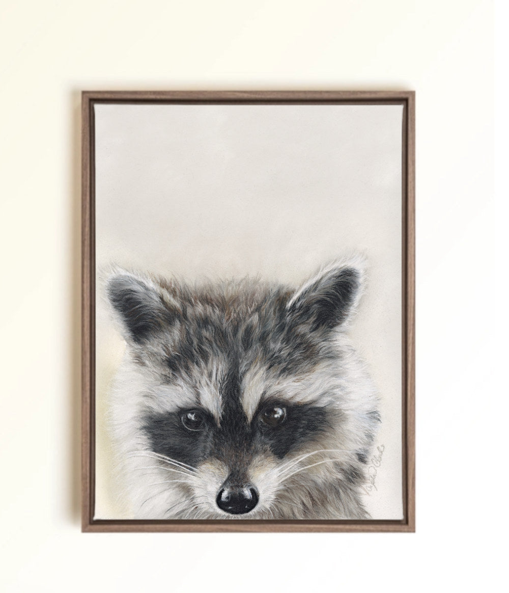 A framed canvas print featuring a detailed portrait of a raccoon with gray, black, and white fur, enclosed in a brown wooden frame against a light beige wall.