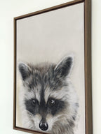 A framed canvas print of a raccoon, featuring detailed gray, black, and white fur with a dark brown wooden frame, hanging on a white wall.